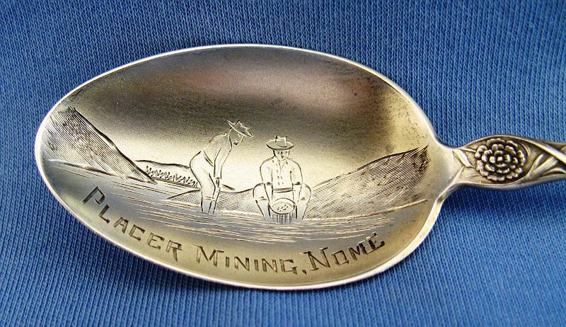 Souvenir Placer Mining Spoon Bowl Nome Alaska.JPG - SOUVENIR MINING SPOON NOME ALASKA - Sterling silver souvenir spoon, engraved mining scene in bowl showing miners panning for gold, marked PLACER MINING, NOME, decorative design with flowers on handle, reverse marked Sterling with makers mark, 5 3/8 in. long  [The Nome mining district is a gold mining district in Alaska. It was discovered in 1898 when Erik Lindblom, Jafet Lindeberg and John Brynteson, the "Three Lucky Swedes", found placer gold deposits on Anvil Creek and on the Snake River, a few miles from the future site of Nome. News of the strike reached the gold fields of the Klondike that winter and by 1899 Anvil City, as the new camp was called, had a population of 10,000. It was not until gold was discovered in the beach sands in 1899 and news reached the outside that the real stampede was on. Thousands poured into Nome during the spring of 1900, as soon as steamships from the ports of Seattle and San Francisco could reach the north through the ice. In the treeless location, tents soon covered the landscape, reaching the water's edge, and extending most of the 30 miles between Cape Rodney and Cape Nome. Buildings of finished board lumber began going up as early as 1899, as soon as ships reached Nome from the states with supplies.  The town was locally known as Anvil City for much of 1899, but the United States Post Office Department insisted on calling the community Nome, apparently because it was thought that a town called Anvil City would be easily confused with the village of Anvik on the lower Yukon. A vote was held and the town’s merchants reluctantly agreed to change the name from Anvil City to Nome.  This was one of the first and was the biggest Alaskan gold rush in North America; only the California and Klondike stampedes were larger. A chaotic and lawless scene ensued, with rampant claim-jumping, crooked judges, and not enough gold found for the 20,000 prospectors, gamblers, shop and saloon-keepers, and prostitutes living in the tent city on the beachfront tundra, at least not at first. Then someone thought to pan the red-and-black streaked beach sands. Within days, gold was found for tens of miles up and down the beach from Nome. More than a million dollars' worth of gold was taken from the beach in 1899. Subsequently the second and third beach lines were discovered and mined. Anvil Creek produced the second-largest gold nugget found in Alaska (182 troy ounces).  Except while prohibited by law during WWII, placer mining near Nome has continued to this day. Over 3.6 million troy ounces of gold have been recovered from the creeks of the Nome District.  A myriad of small hard-rock gold deposits were exploited near Nome, but production was very small, compared to the placer deposits, and none of the hard rock mines operated for more than a few years.]